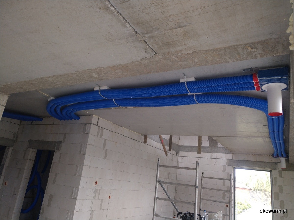 Niebieskie rury wentylacyjne rekuperacji zamontowane pod betonowym stropem w surowym wnętrzu budynku z białych bloczków, widoczna drabina i otwór okienny.