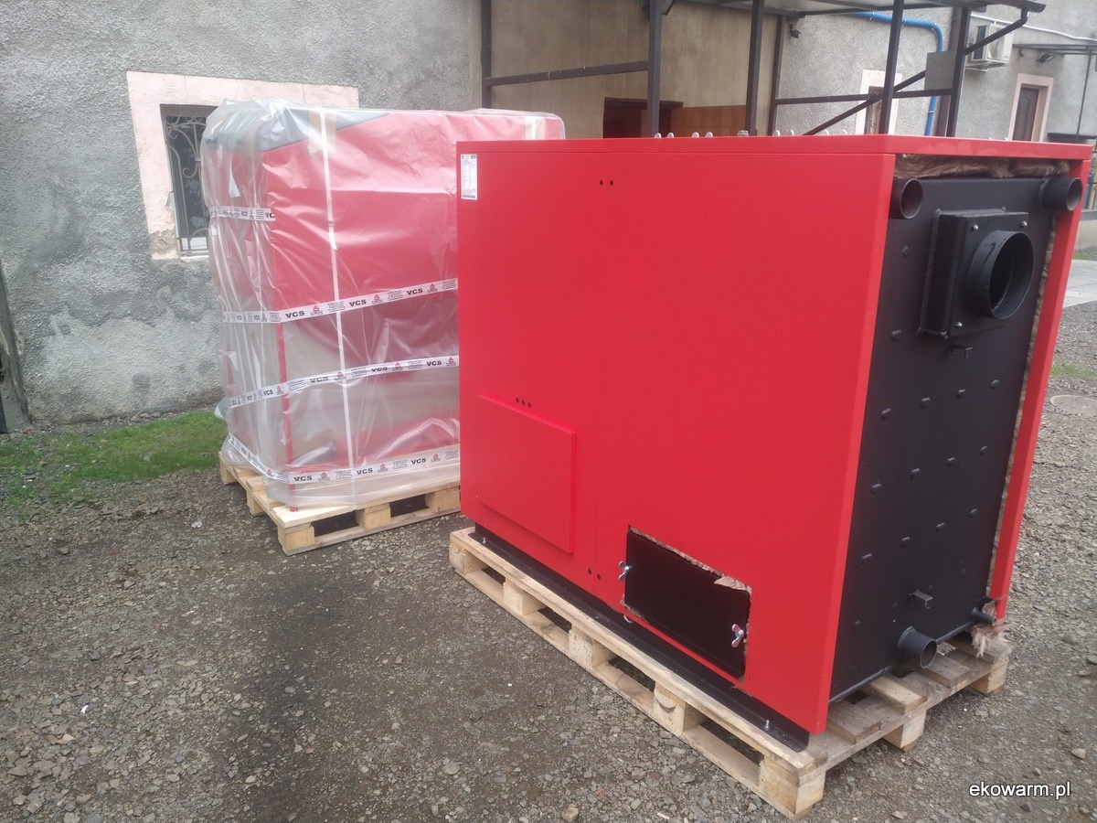 Dwa kotły stałopalne na paletach przed budynkiem. Jeden czerwony, drugi zapakowany w folię z napisem VCS.