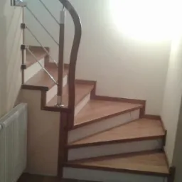 Klatka schodowa z drewnianymi stopniami i metalową balustradą z drewnianym poręczą, widoczny grzejnik pod schodami.