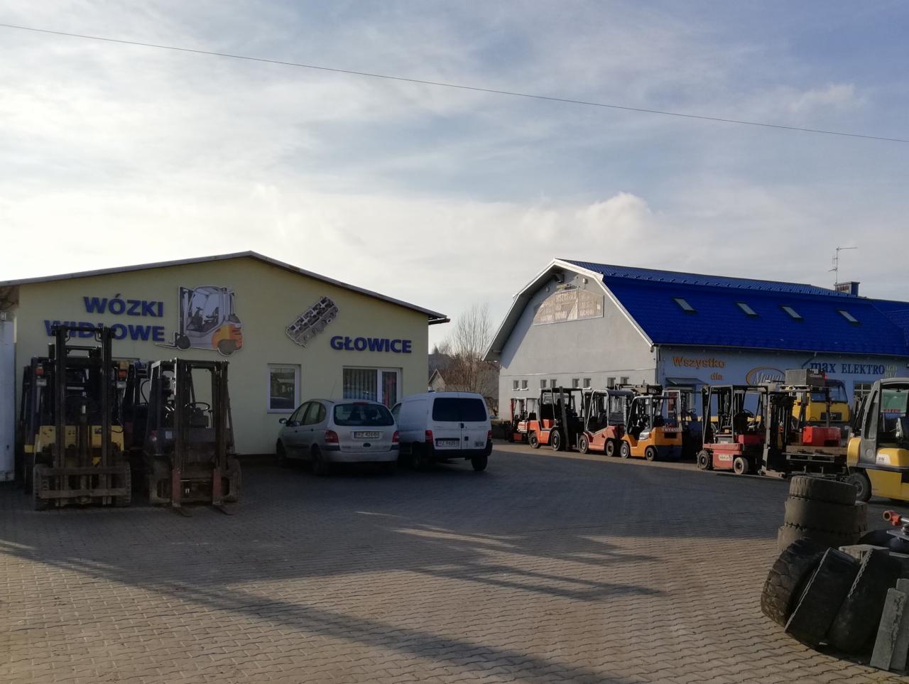 Plac firmy w Tyczynie z zaparkowanymi wózkami widłowymi różnych marek i kolorów, budynek z napisem 'Wózki Widłowe Głowiece' oraz budynek z niebieskim dachem