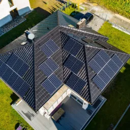 Widok z góry na dom z ciemną dachówką i panelami słonecznymi rozmieszczonymi w kilku sekcjach dachu, z widocznym tarasem i ogrodem.