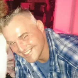 Uśmiechnięty mężczyzna w niebieskiej koszuli w kratę, z bliska, w tle rozmazane postacie i czerwone oświetlenie.