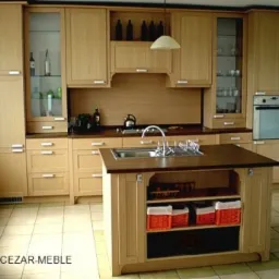 Jasna, drewniana kuchnia na wymiar z wyspą, ciemny blat, przeszklone szafki wiszące, oświetlenie punktowe, widoczne butelki i akcesoria kuchenne.