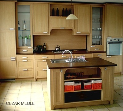 Jasna, drewniana kuchnia na wymiar z wyspą, ciemny blat, przeszklone szafki wiszące, oświetlenie punktowe, widoczne butelki i akcesoria kuchenne.