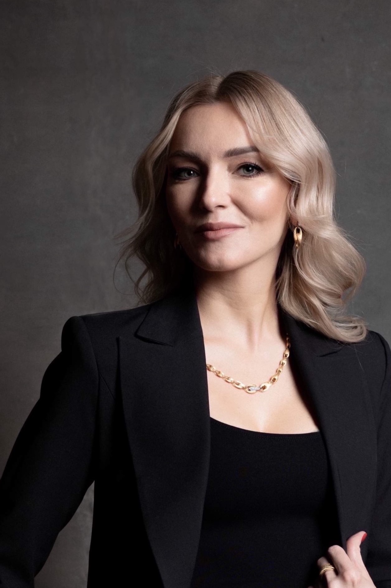 Portret kobiety w czarnym żakiecie i złotej biżuterii na tle szarej ściany.