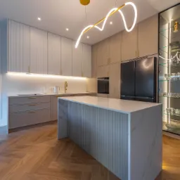 Nowoczesna kuchnia w odcieniach beżu z wyspą i marmurowym blatem. Oświetlenie LED podkreśla minimalistyczny design i szklaną witrynę. Parkiet w jodełkę.