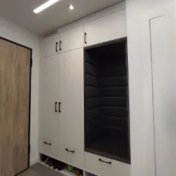 Biała szafa wnękowa z czarnymi uchwytami, wbudowanym siedziskiem obitym ciemną tapicerką i otwartymi półkami na buty, widok na fragment drzwi z czarną ramą.