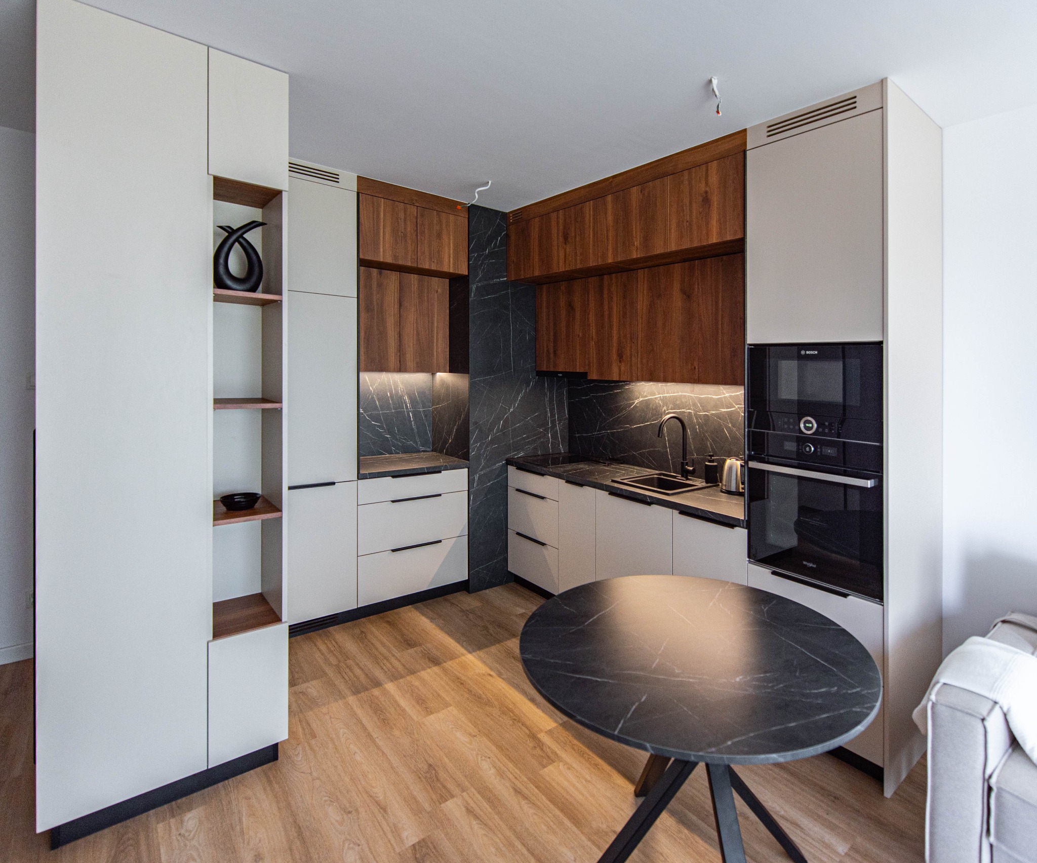 Nowoczesna kuchnia z białymi i drewnianymi szafkami, czarnym blatem i okrągłym stołem z blatem imitującym marmur.