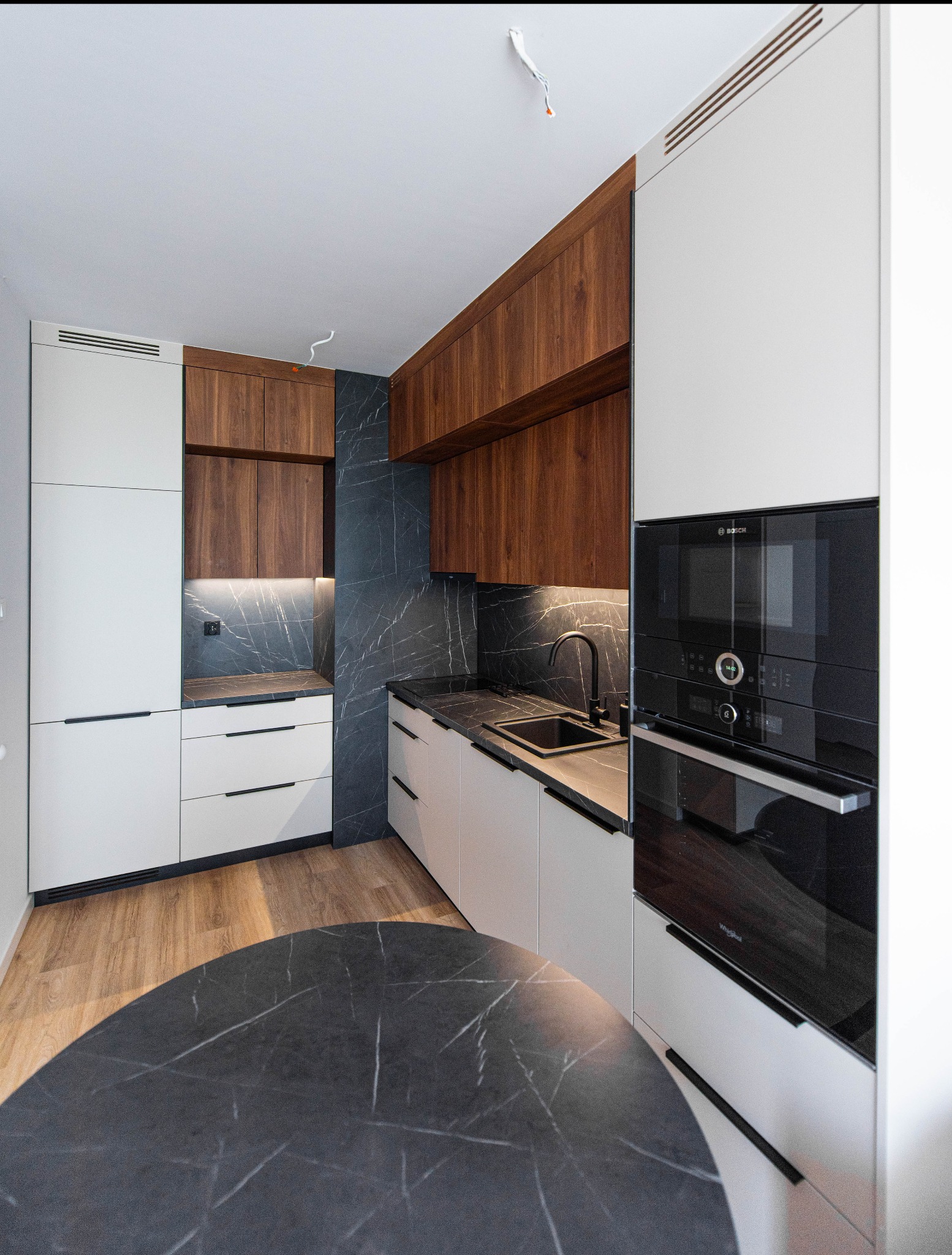 Nowoczesna kuchnia z białymi frontami i drewnianymi szafkami górnymi, czarny blat z marmurowym wzorem, okrągły stół z blatem z tego samego materiału, czarna armatura i sprzęt AGD.