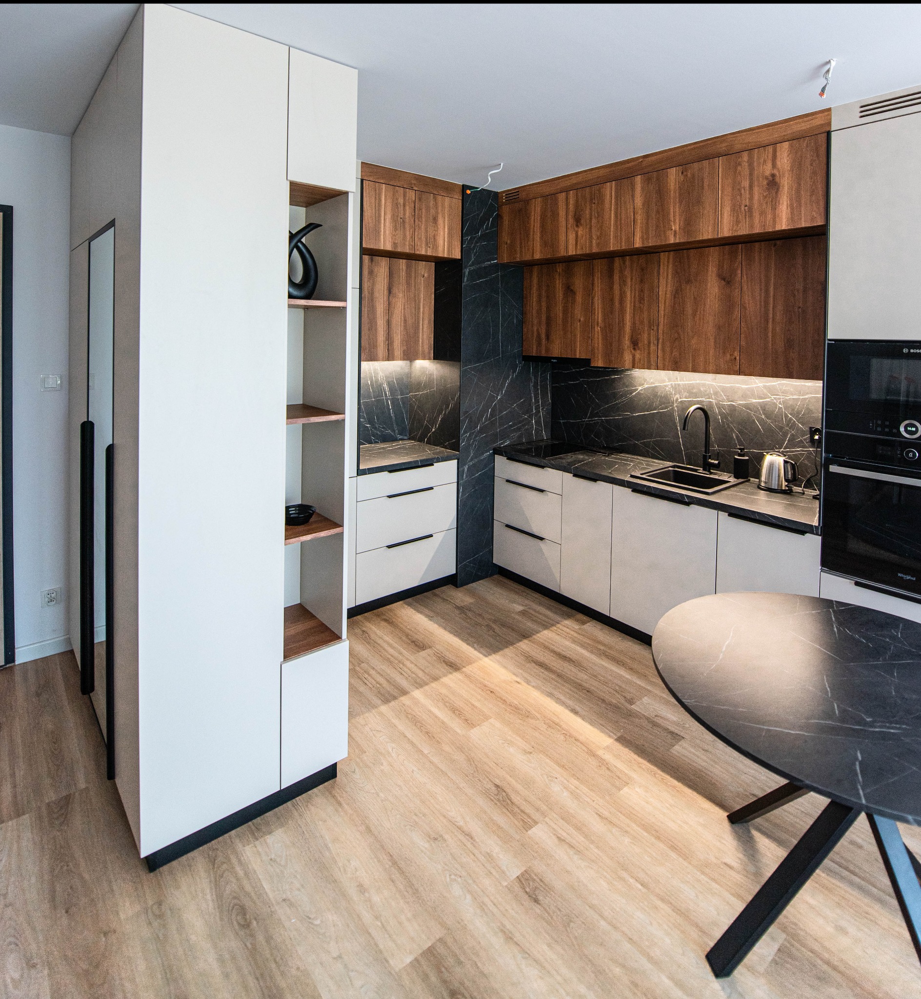 Nowoczesna kuchnia z drewnianymi szafkami górnymi, białymi dolnymi, blatem i ścianą z czarnego marmuru, okrągłym stołem z czarnym blatem i czarnymi nogami oraz półkami z dekoracjami.