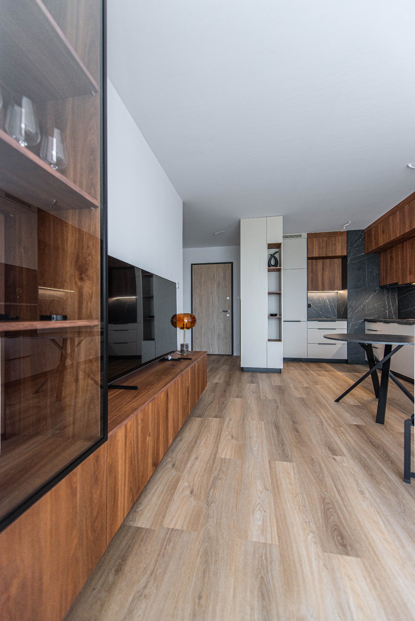 Wnętrze mieszkania z drewnianą zabudową kuchenną, szafkami wiszącymi i blatem z imitacją marmuru, widok na szklaną witrynę i drewnianą podłogę.