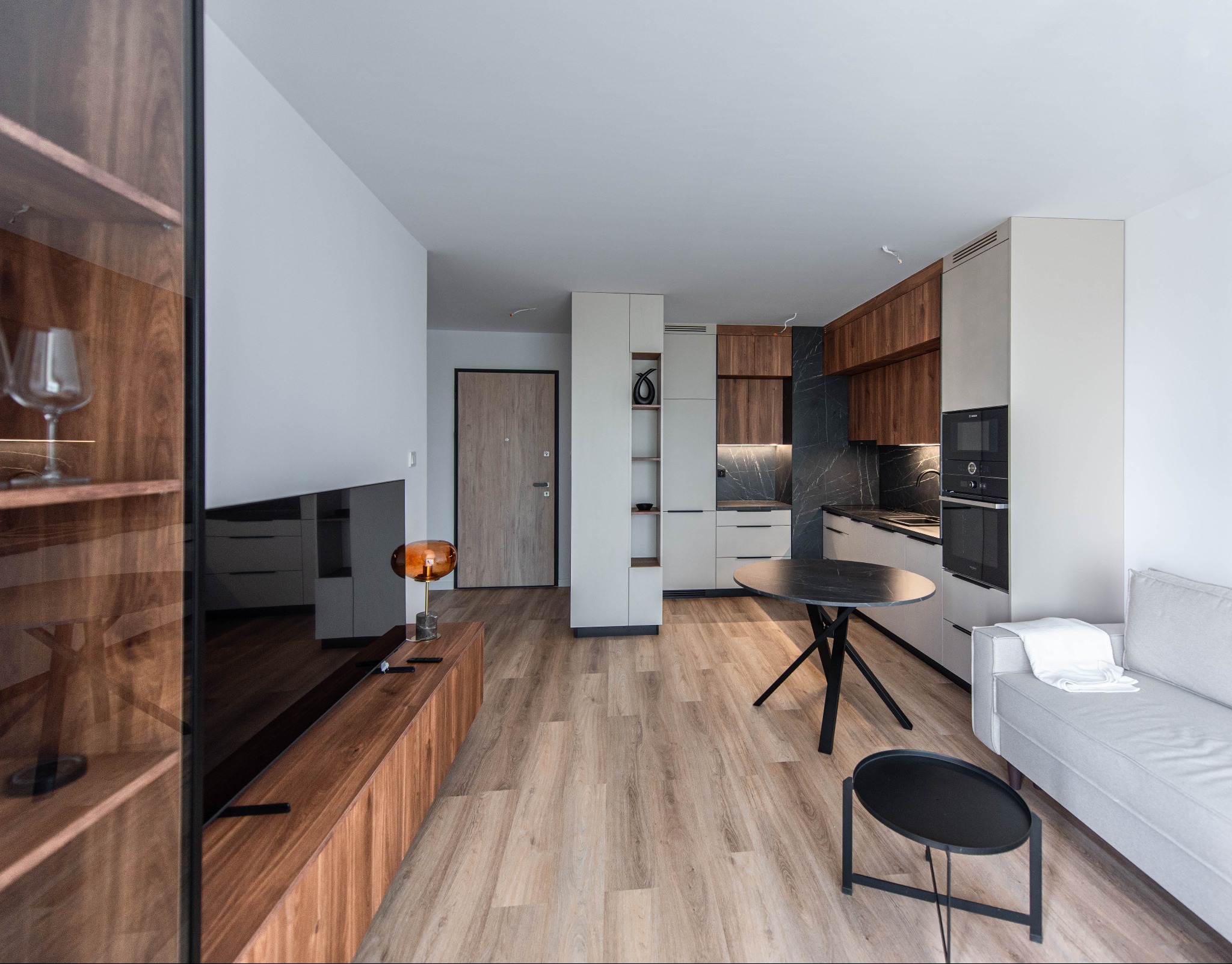 Nowoczesne wnętrze apartamentu z otwartą kuchnią, drewnianymi akcentami i marmurowym blatem, oświetleniem LED pod szafkami, okrągłym stołem z czarnym blatem i szarą sofą.