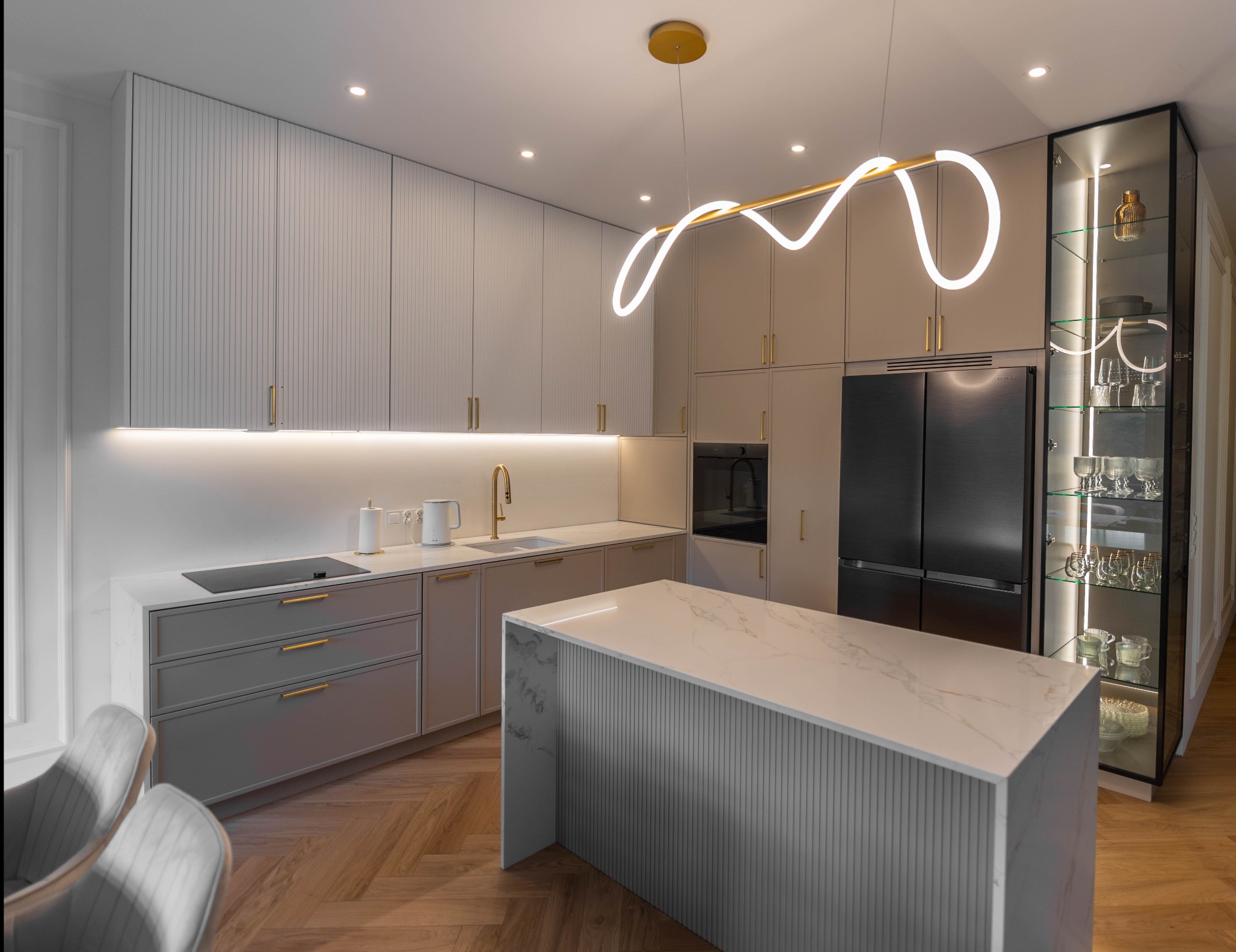 Nowoczesna kuchnia z wyspą, fronty szafek z pionowymi żłobieniami, złote uchwyty i armatura, podświetlenie LED, szklana witryna z zastawą.