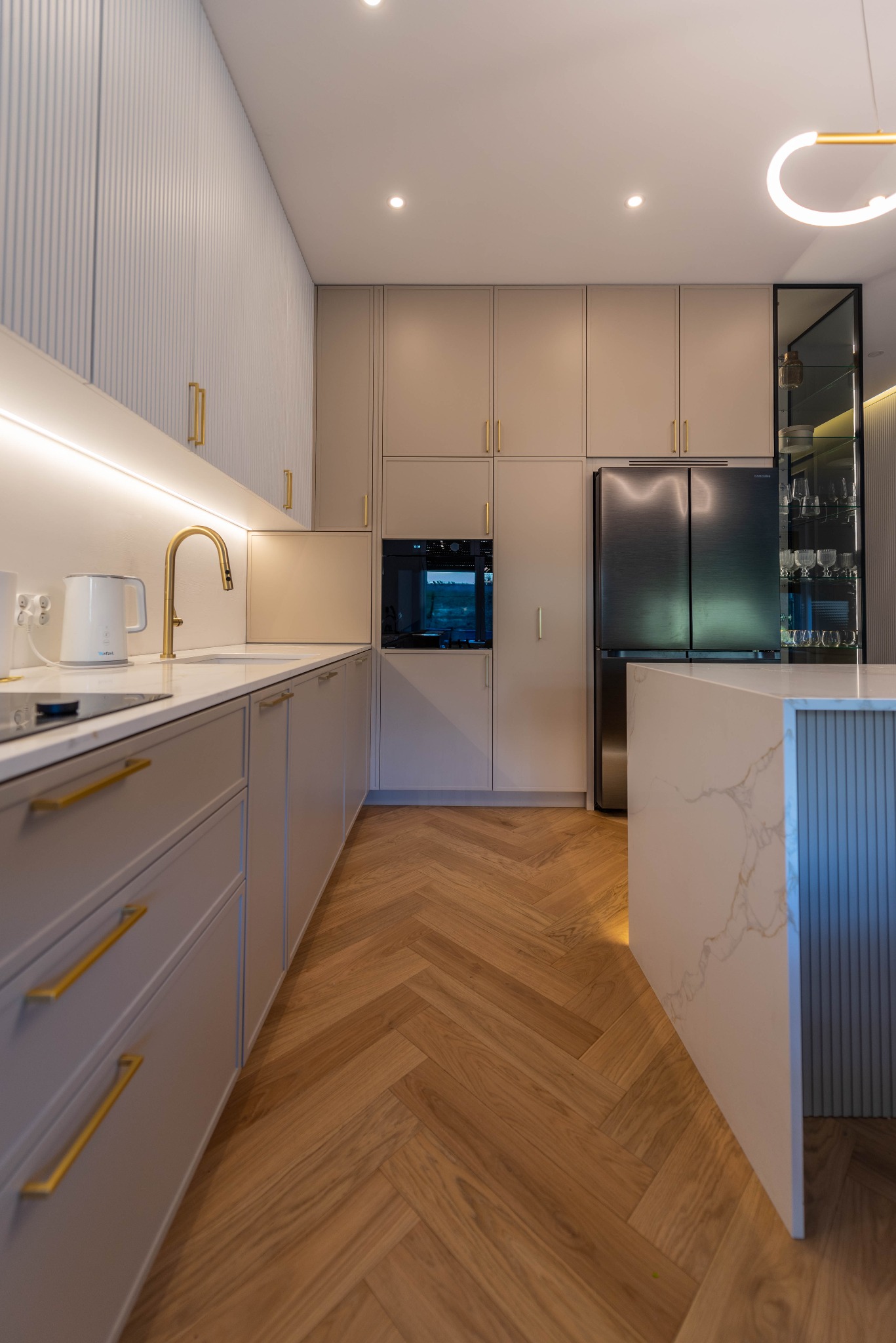 Nowoczesna kuchnia z szarymi szafkami, złotymi uchwytami i armaturą, blatem z efektem marmuru, drewnianą podłogą ułożoną w jodełkę oraz wyspą kuchenną z podświetleniem.