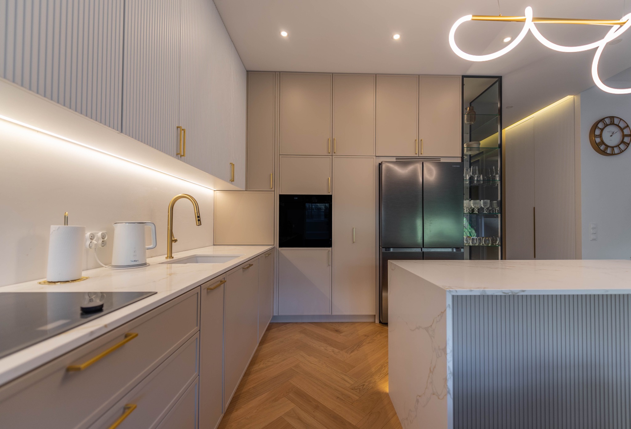 Nowoczesna kuchnia w beżach i bieli z drewnianą podłogą w jodełkę, złotymi akcentami, wyspą z blatem imitującym marmur i dekoracyjnym oświetleniem.