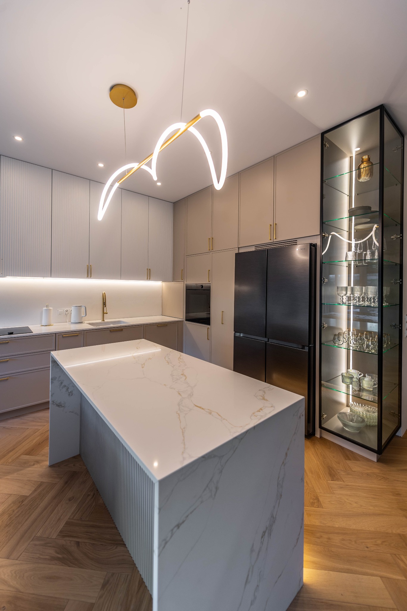 Elegancka kuchnia: wyspa z blatem marmurowym, szafki w odcieniach beżu, oświetlona witryna z szkła i czarnych ram, parkiet w jodełkę i nowoczesna lampa.