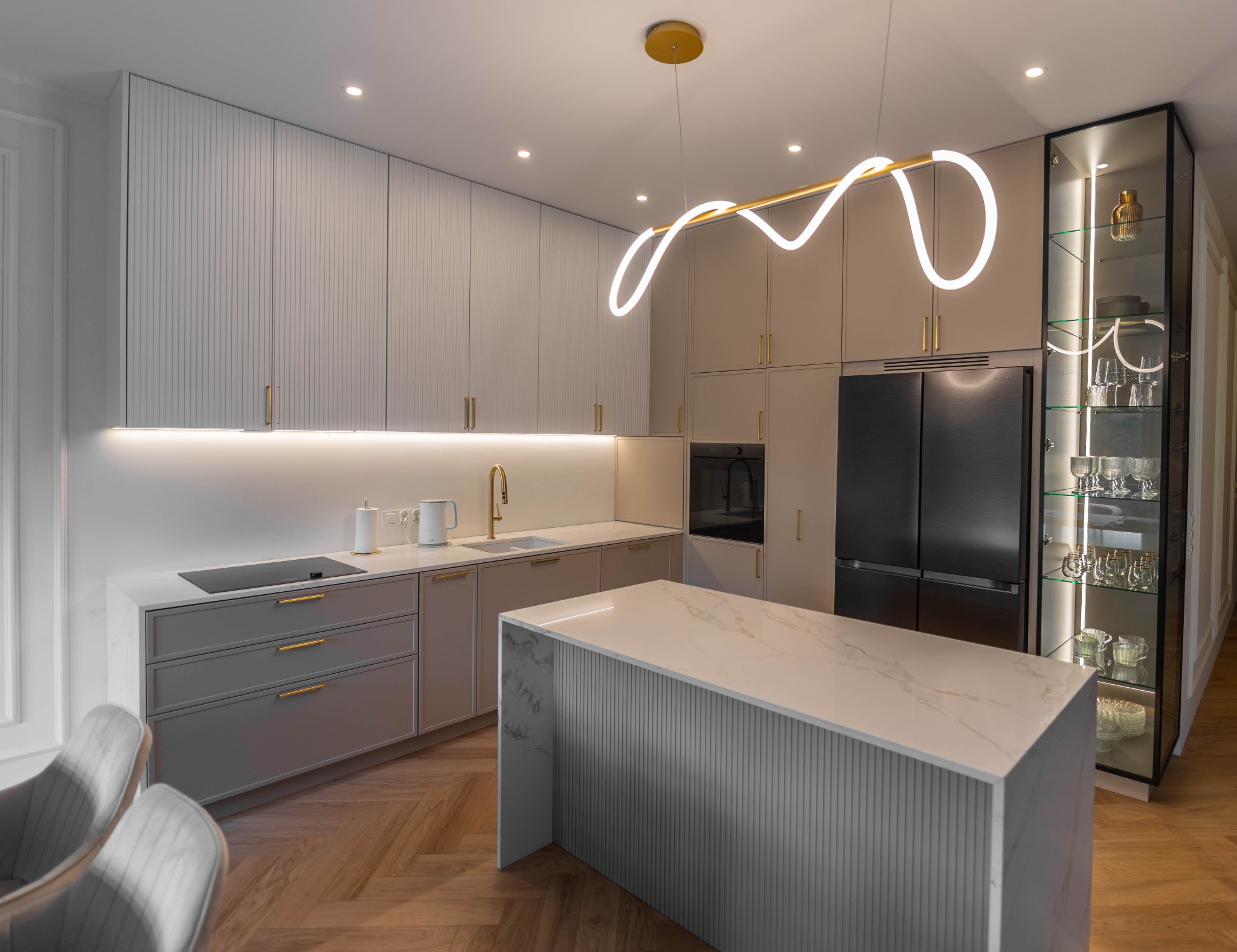Nowoczesna kuchnia w odcieniach szarości z wyspą, złotymi akcentami i oświetleniem LED. Szafka z przeszkleniami eksponuje dekoracyjne naczynia.