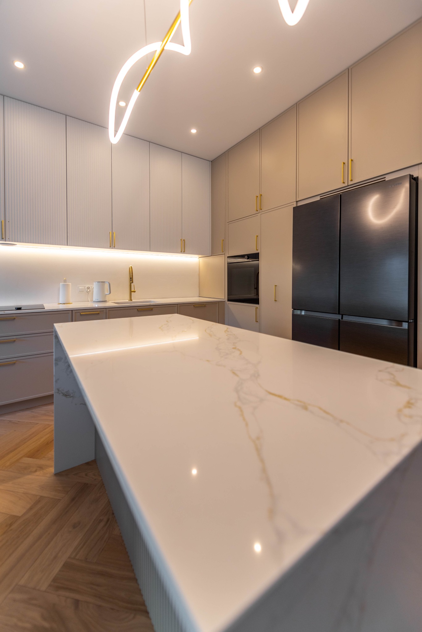 Beżowa kuchnia z wyspą i złotymi akcentami: szafki z frezowanymi frontami, marmurowy blat z podświetleniem LED, podłoga w jodełkę, nowoczesna lampa.