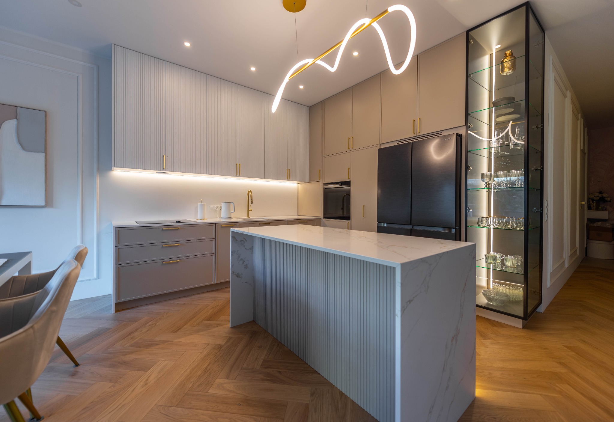 Nowoczesna kuchnia w odcieniach beżu z wyspą i marmurowym blatem. Oświetlenie LED podkreśla minimalistyczny design i szklaną witrynę. Parkiet w jodełkę.