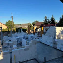 Budowa domu jednorodzinnego: wznoszone ściany z białych bloczków, widoczne palety z materiałem, szalunki kolumn, oraz elementy zbrojenia, na tle zabudowy jednorodzinnej w pogodny dzień.