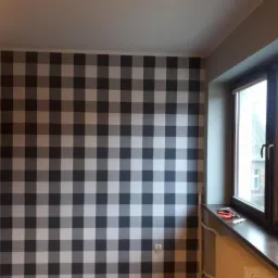 Ściana w pokoju wykończona tapetą w dużą szarą kratę, widok na okno z ciemną ramą, na parapecie narzędzia i grzejnik pod oknem.