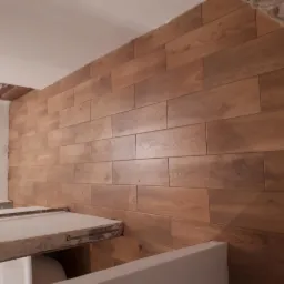 Ściana wyłożona płytkami imitującymi drewno, ułożonymi poziomo, z widocznym fragmentem sufitu w trakcie prac wykończeniowych i fragmentem zabudowy w dolnej części kadru.