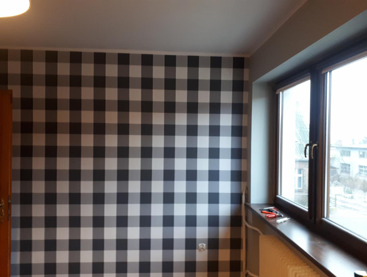 Ściana w pokoju wykończona tapetą w dużą szarą kratę, widok na okno z ciemną ramą, na parapecie narzędzia i grzejnik pod oknem.