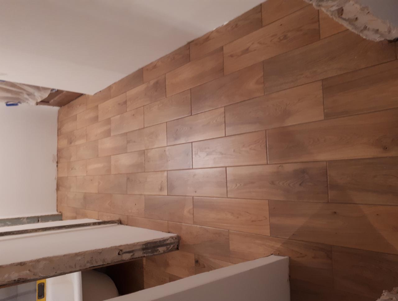 Ściana wyłożona płytkami imitującymi drewno, ułożonymi poziomo, z widocznym fragmentem sufitu w trakcie prac wykończeniowych i fragmentem zabudowy w dolnej części kadru.