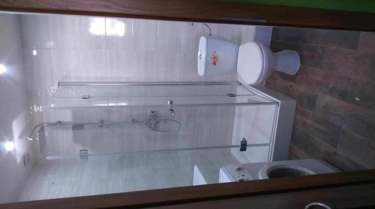 Nowoczesna łazienka po remoncie, widok na kabinę prysznicową z przezroczystego szkła, toaletę marki Koło i pralkę, na podłodze ciemne panele imitujące drewno, a na ścianach jasne płytki.