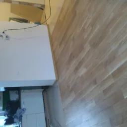 Nowo ułożona podłoga z paneli w jasnym odcieniu drewna, widoczna z perspektywy sufitu, z fragmentem białej ściany i wyposażenia kuchni w tle.