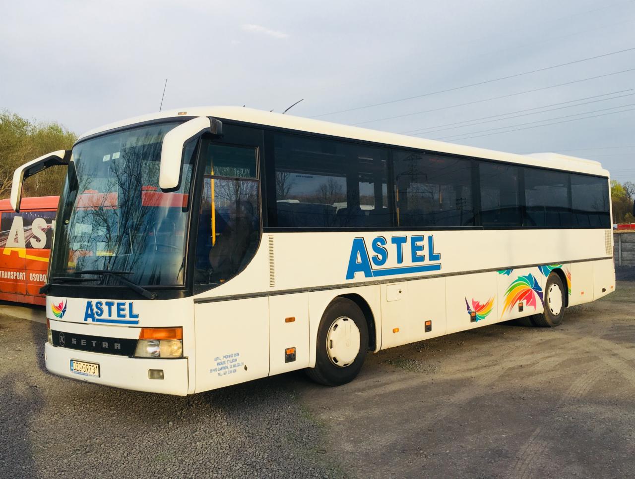 Autokar firmy Astel z Wrocławia, z otwartymi drzwiami, stojący na żwirowym parkingu, z kolorowym motywem piór na boku.