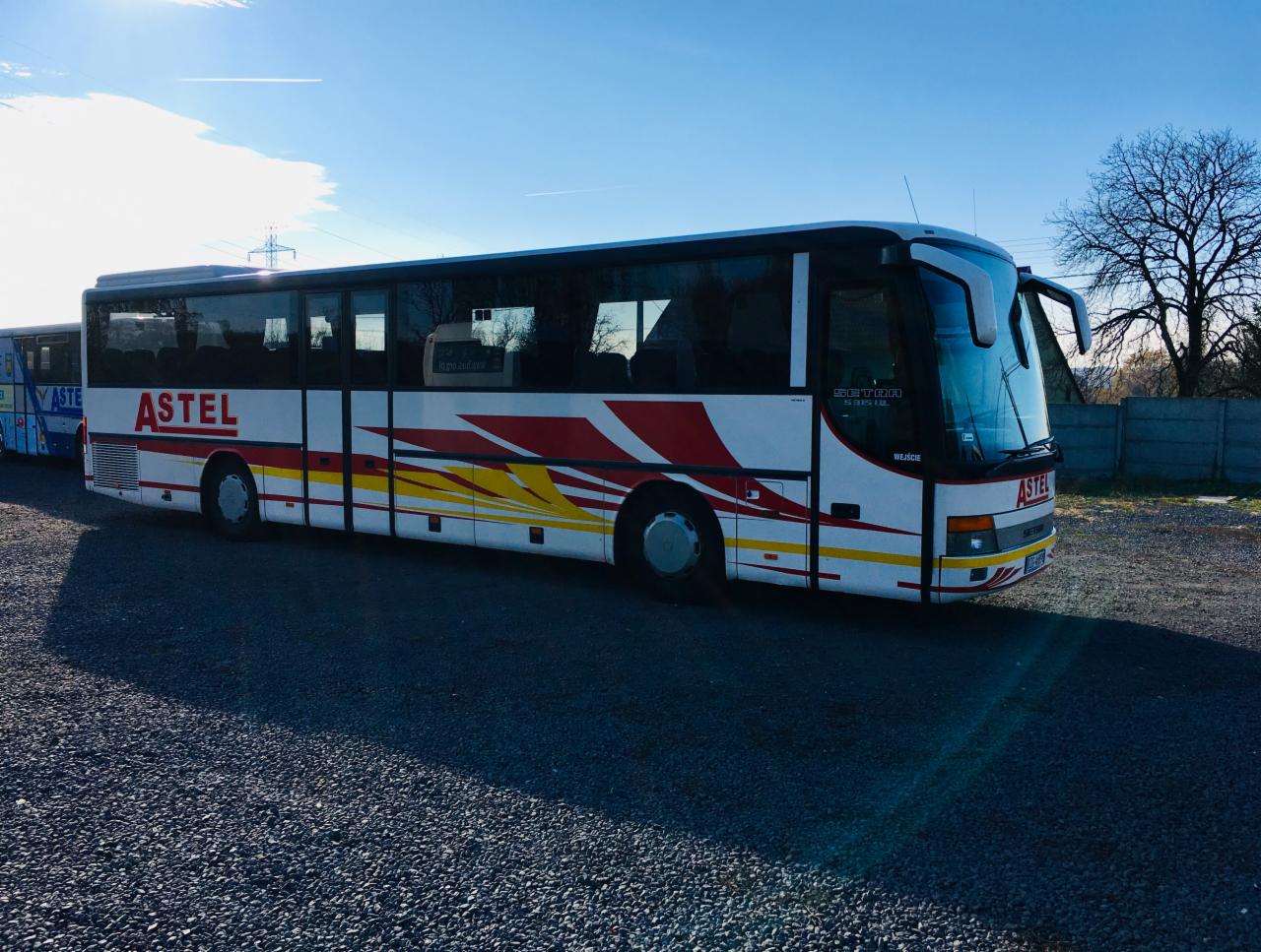 Autokar marki Setra z nazwą firmy Astel na boku, stojący na żwirowym parkingu w słoneczny dzień.