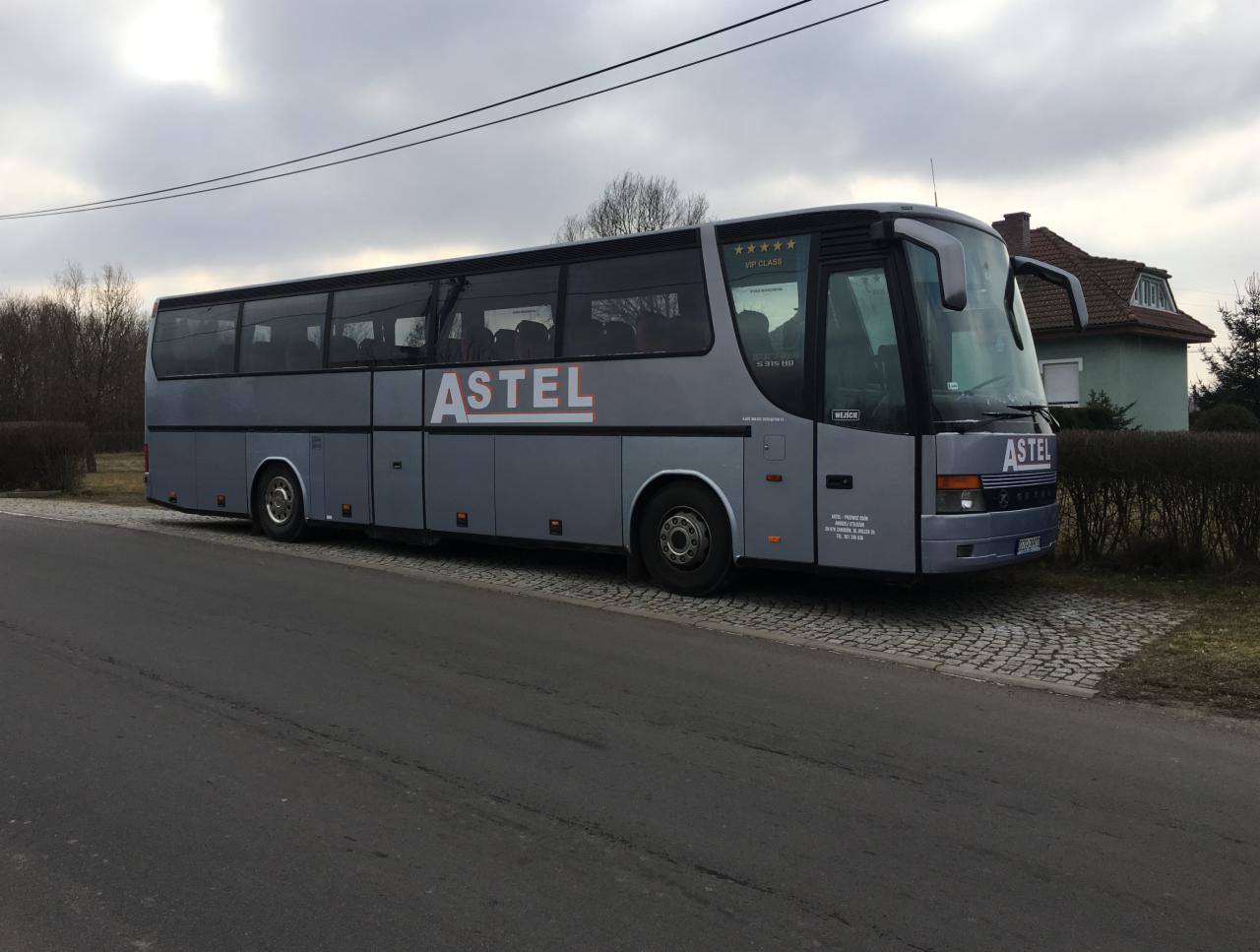 Szary autokar firmy ASTEL z napisem VIP CLASS i pięcioma gwiazdkami, zaparkowany na kostce brukowej przy drodze i zielonym budynku w tle.
