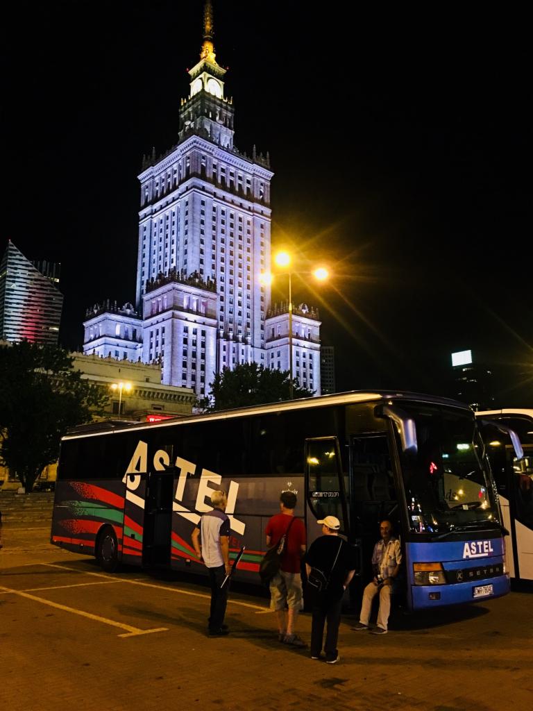 Autokar firmy Astel na tle nocnego Pałacu Kultury w Warszawie, pasażerowie wsiadają.