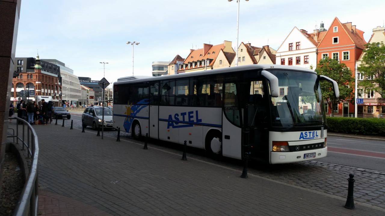 Biały autobus firmy Astel z otwartymi drzwiami stoi na chodniku w centrum miasta, na tle kamienic ze spadzistymi dachami.