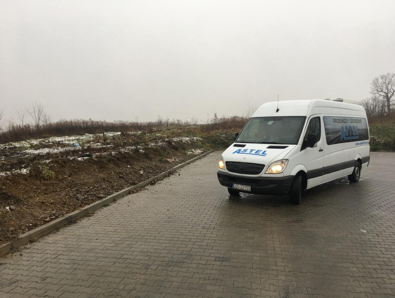 Biały bus z logo firmy przewozowej Astel na tle pochmurnego krajobrazu z fragmentami śniegu.