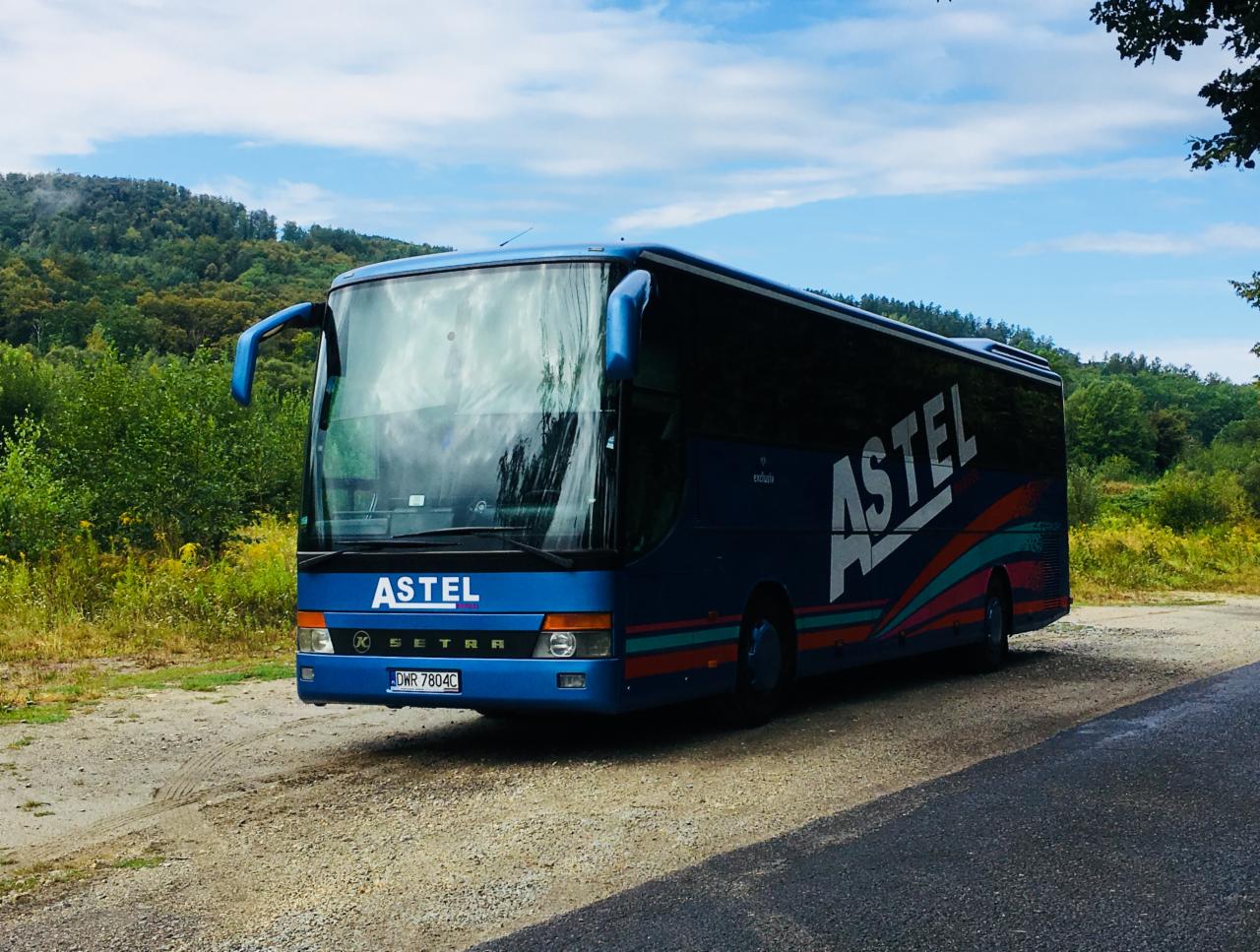 Niebieski autokar Setra marki Astel z kolorowym logo na tle zieleni i błękitnego nieba, zaparkowany na żwirowej drodze.