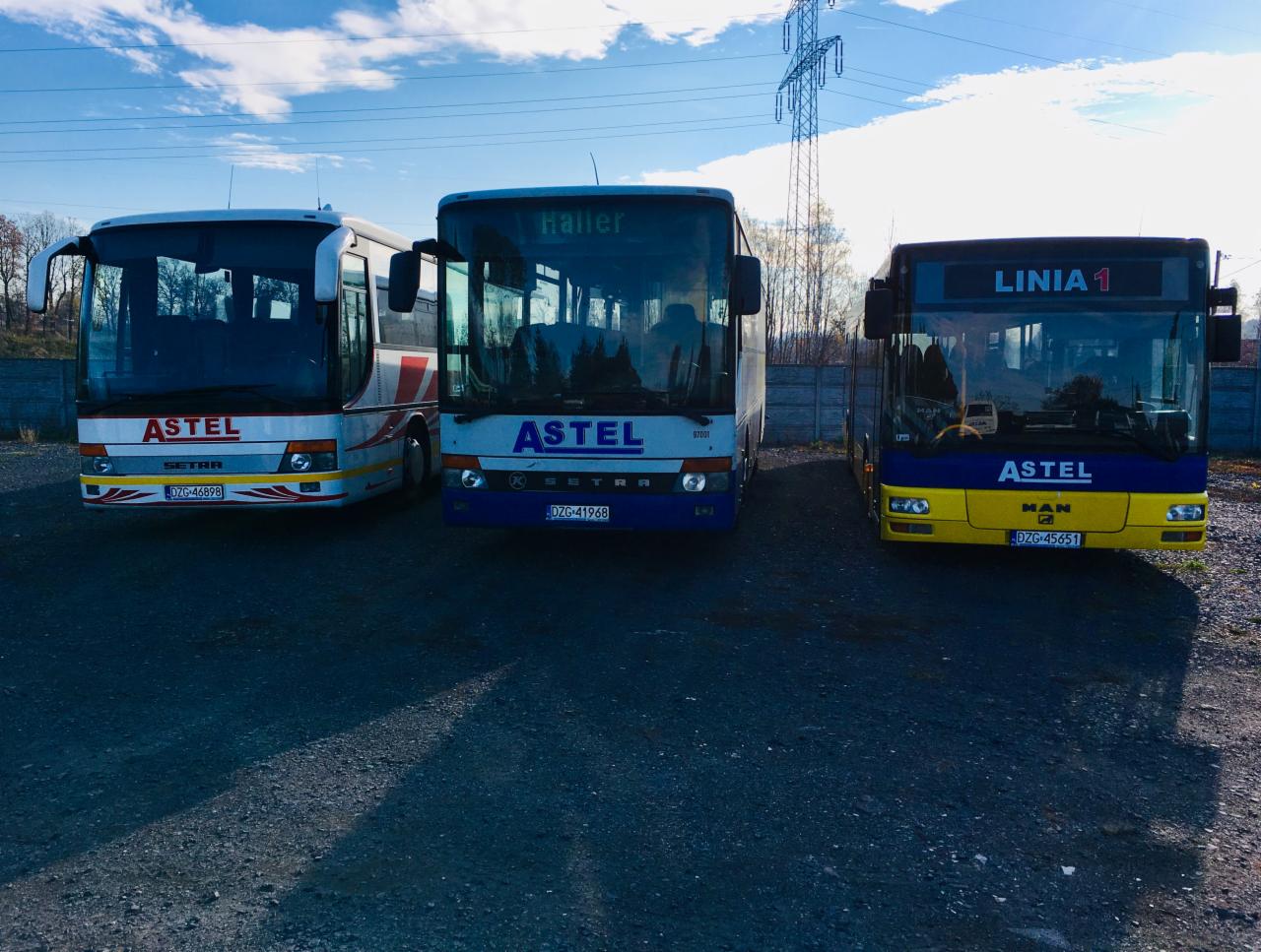 Trzy autobusy firmy Astel, różnych modeli i malowań, zaparkowane na żwirowym placu z betonowym ogrodzeniem w tle, widoczne linie wysokiego napięcia.