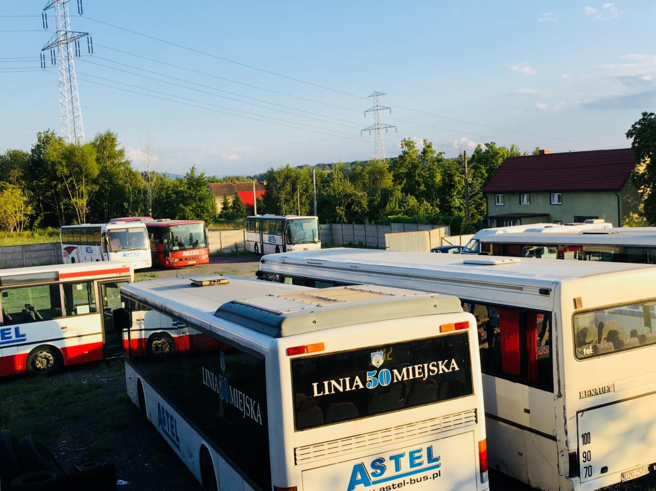 Flota autobusów miejskich firmy Astel na parkingu, widoczne linie autobusowe z numerem 50, słup wysokiego napięcia w tle.