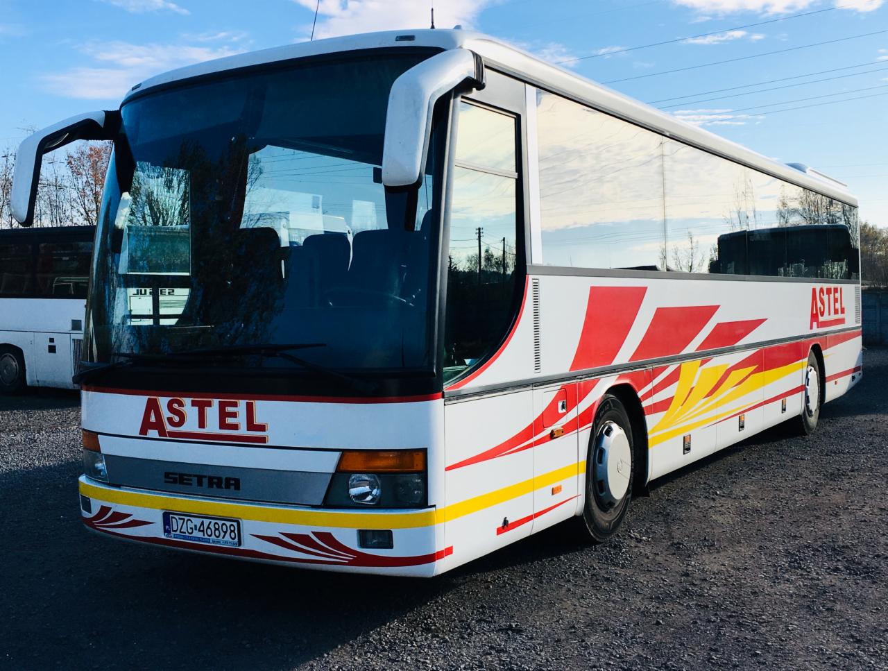 Biały autokar marki Setra z czerwono-żółtymi elementami graficznymi i logo firmy Astel, zaparkowany na żwirowym parkingu w słoneczny dzień.