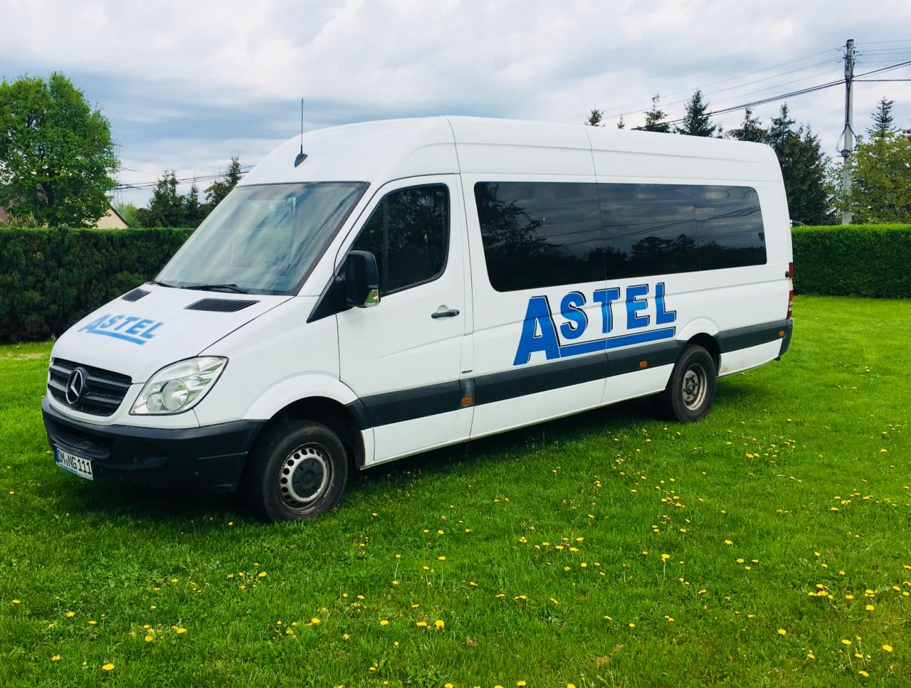 Biały bus Mercedes Sprinter z niebieskim logo 'ASTEL' na zielonej trawie z mleczami, pochmurne niebo w tle.