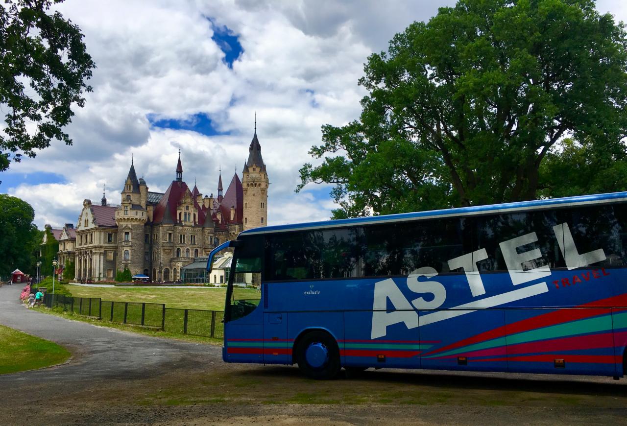 Niebieski autokar turystyczny z logo Astel Travel na tle zamku w Mosznej i zieleni drzew.