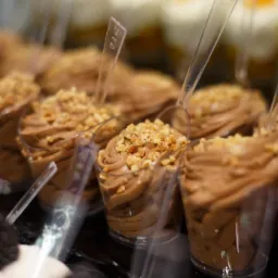 Porcja musu czekoladowego w przezroczystym kubeczku, posypana posiekanymi orzechami, widoczna w witrynie obok innych deserów.