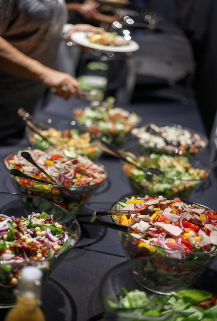Bufet sałatkowy z różnorodnymi sałatkami w szklanych misach, osoba nakłada sałatkę na talerz w tle, ciemne tło.