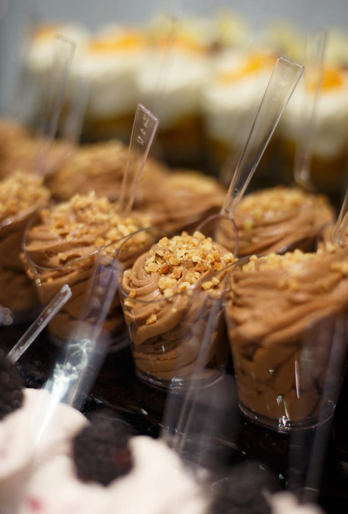 Porcja musu czekoladowego w przezroczystym kubeczku, posypana posiekanymi orzechami, widoczna w witrynie obok innych deserów.