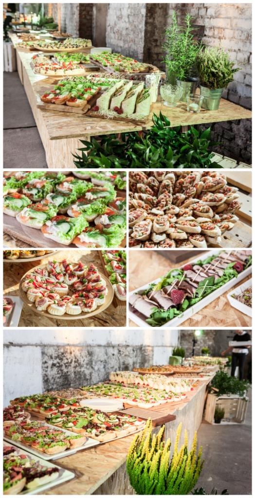 Długi stół z przekąskami cateringowymi: kanapki, tortille, koreczki i sałatki, udekorowany zielenią, ustawiony w surowym wnętrzu ceglanego budynku.