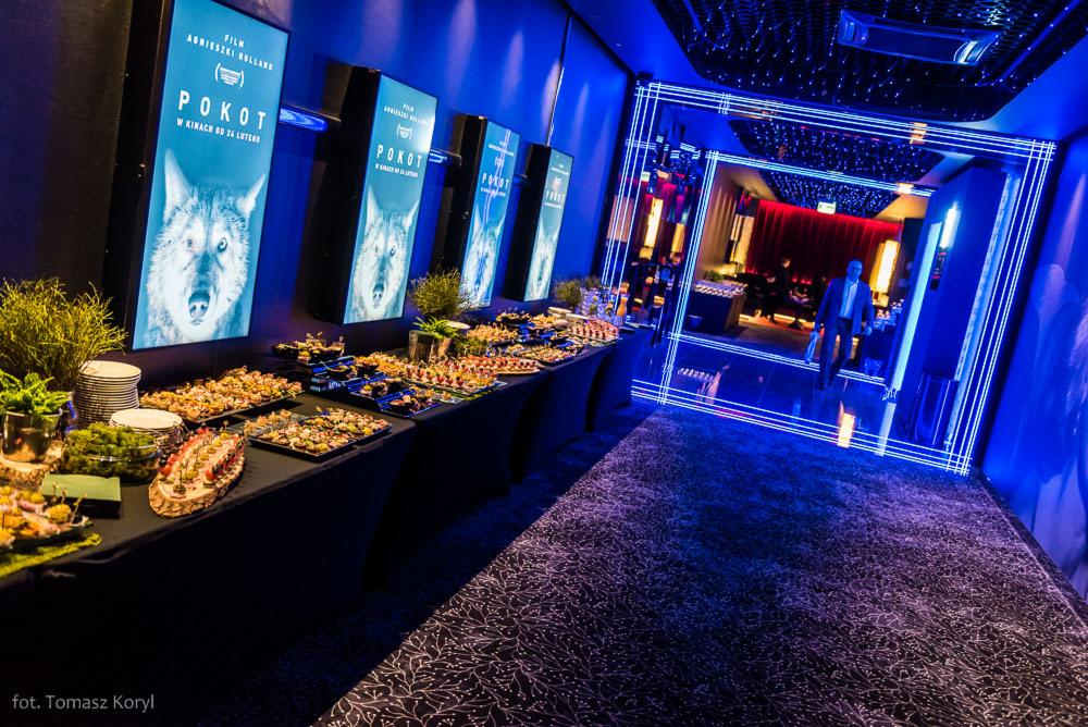Długi stół z czarnym obrusem uginający się pod ciężarem przekąsek, kanapek i przystawek na tle plakatów filmowych i futurystycznego oświetlenia.