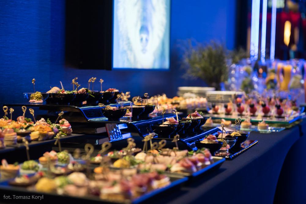 Stół cateringowy z różnorodnymi przekąskami na czarnych tacach, podświetlony niebieskim światłem, w tle dekoracja ścienna i stos talerzy.