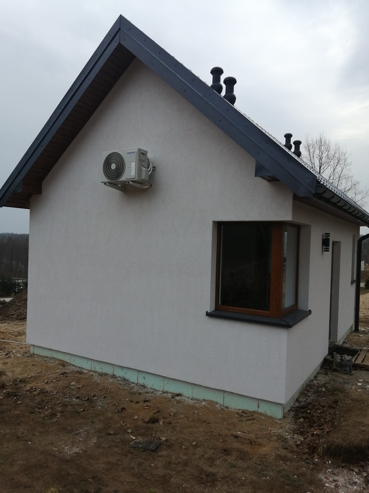 Zewnętrzny klimatyzator Samsung zamontowany na ścianie nowo otynkowanego domu z grafitowym dachem i drewnianą stolarką okienną, widok z boku w pochmurny dzień.