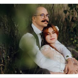 OTO STUDIO Sp. z o.o. - Para narzeczonych w objęciach siedzi pod drzewem: mężczyzna z wąsami w okularach i kamizelce, kobieta w białej sukni z kwiatem we włosach, na ramieniu kobiety widoczny tatuaż półksiężyca.